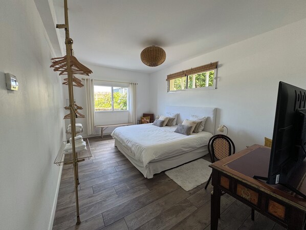 A bedroom with a large bed, a desk, a chair, a TV, a window with a view of greenery, a hanging light fixture, and a bench by the window.