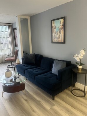 Un salón con un sofá azul oscuro, una mesa de centro de vidrio, un cuadro enmarcado en la pared y una mesita auxiliar con una orquídea blanca.