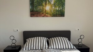 A bedroom with a bed, two nightstands, a wall-mounted painting of a forest, and a power outlet on each nightstand.