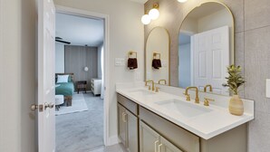 A modern bathroom with a double sink, gold fixtures, and a large mirror with three lights. - South Austin Luxe Boho House 4BR 2.5BA With SPA, Pool & EV Charger (Mustang Ridge)