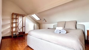 Une chambre avec un grand lit, une lucarne, un plancher en bois et une étagère intégrée où sont rangées des chaussures.