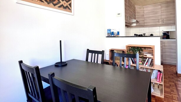 Ein Essbereich mit einem dunklen Holztisch und -stühlen, einem gemusterten Wandbehang und Blick in eine Küche mit Holzausbau und Arbeitsplatte.