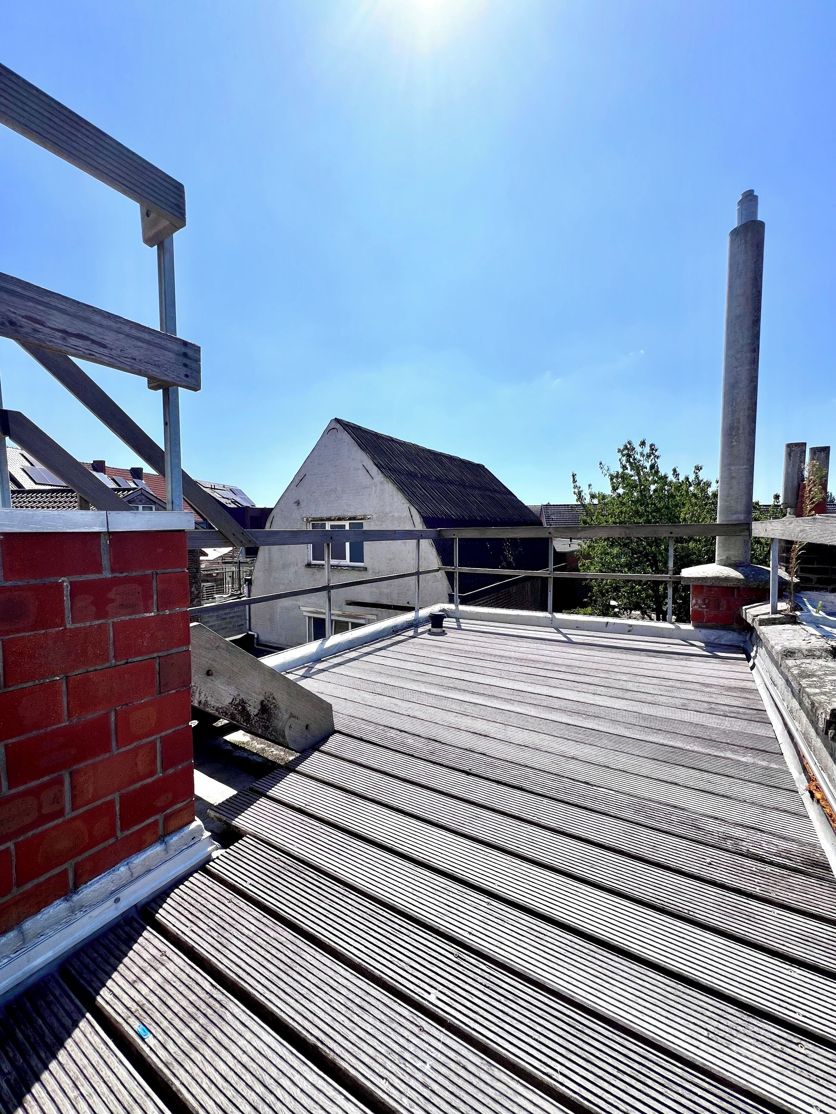Una terraza en la azotea con piso de madera, un muro de ladrillo y un edificio con techo inclinado al fondo.