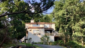 A two-story house with a balcony, surrounded by dense trees and a gravel driveway. - Scenic & Serene: 3BR/3BA w Panoramic Hudson Views! A Birder's paradise (Croton-on-Hudson)