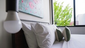 Un lit bien fait, avec une literie blanche et des coussins verts et blancs, un tableau accroché au mur et une fenêtre donnant sur des arbres.