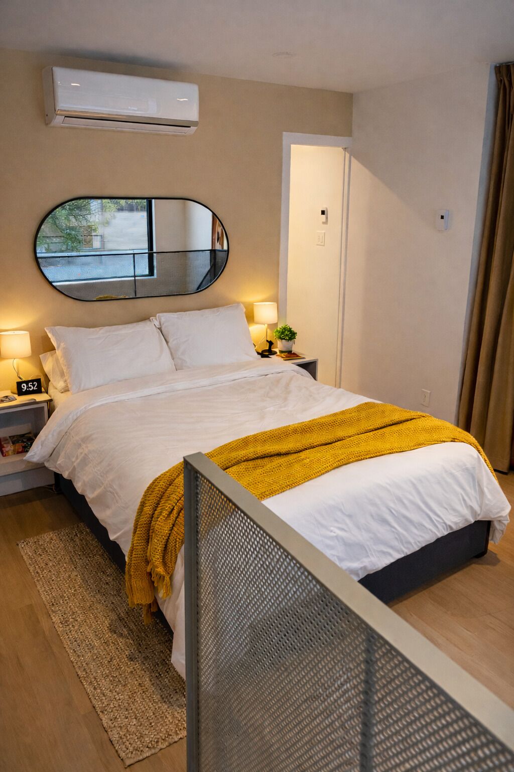 A neatly made bed with white linens and a yellow throw blanket, a round mirror on the wall, a bedside table with a lamp, and an air conditioning unit above the bed.