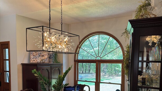 A dining area with a wooden table set for four, surrounded by chairs. A chandelier hangs above, and a large arched window provides a view of the outdoors.