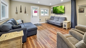 Sala de estar con un sofá gris, un sillón gris y una mesa de centro de madera. Hay un televisor montado en la pared y una pequeña mesita auxiliar.