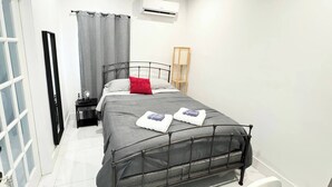 Un dormitorio moderno con estructura de cama metálica, cubrecama gris y dos almohadas. Hay una mesita de noche con lámpara, una ventana con cortinas y un equipo de aire acondicionado en la pared.