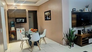 A dining area with a glass table and white chairs, a water dispenser, a small refrigerator, and a television on a wooden stand.