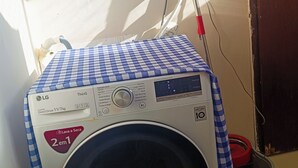 A front-loading washing machine with a blue and white checkered cloth on top, a red mop leaning against the wall, and a wooden door in the background.