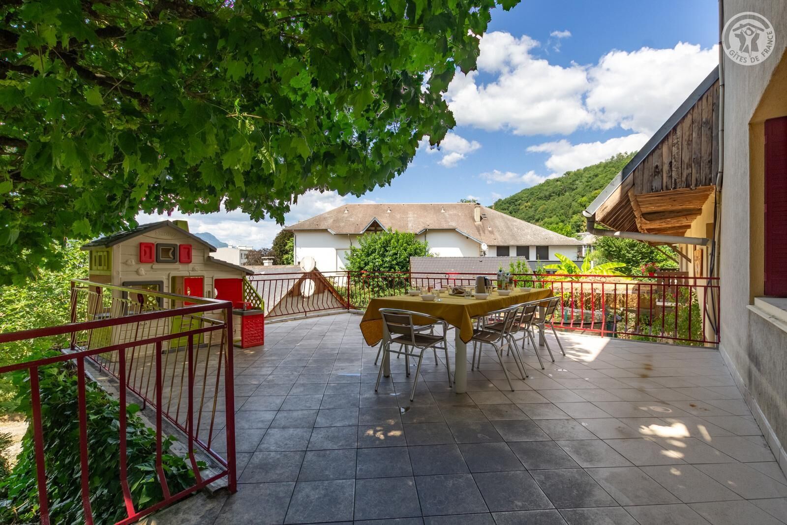 A patio with a table and chairs, surrounded by a red fence, with buildings and greenery in the background.