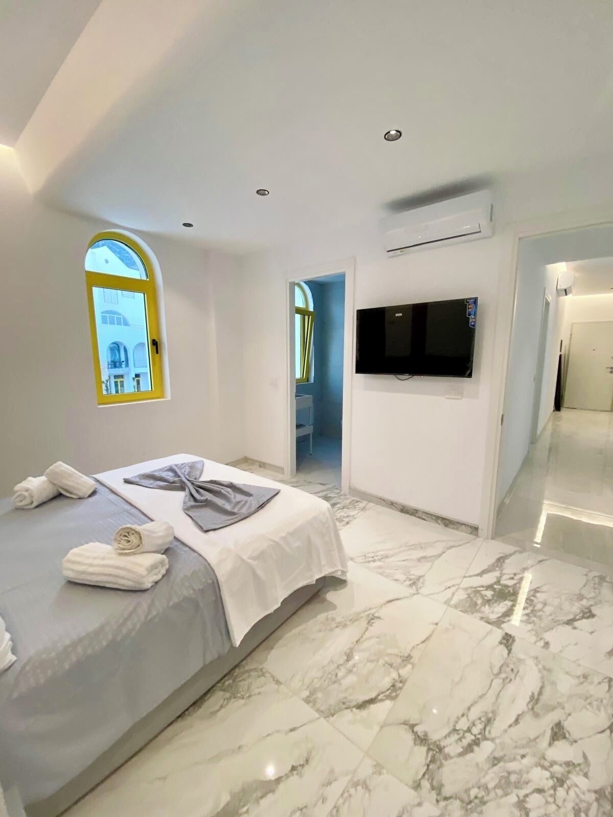 Quarto moderno com cama, toalhas, televisão e banheiro visível por um arco.