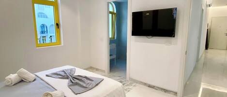 Quarto moderno com cama, toalhas, televisão e banheiro visível por um arco.