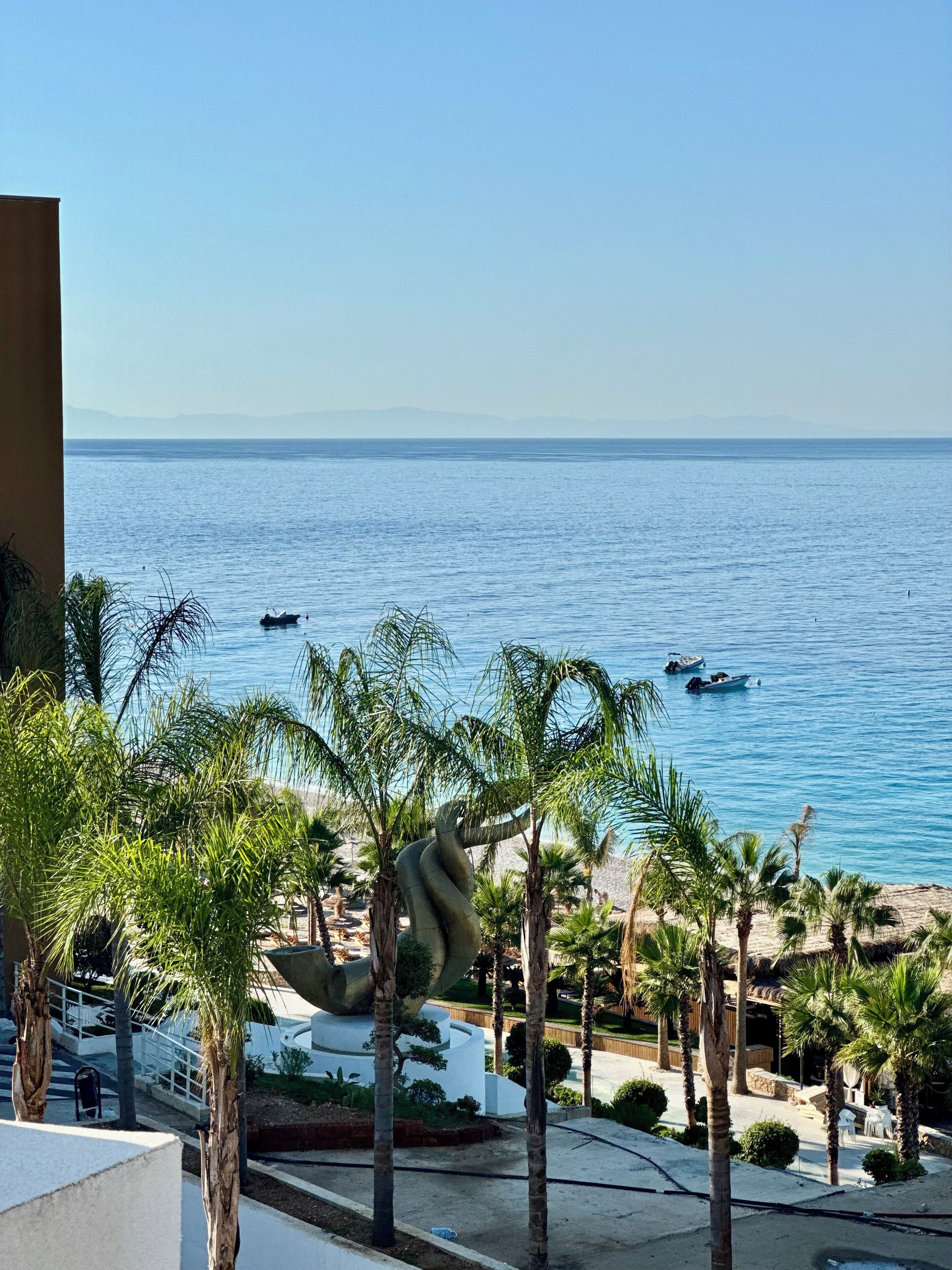 Blick auf einen Strand mit Palmen und einer großen Skulptur im Vordergrund, mit Blick auf das Meer und Boote.