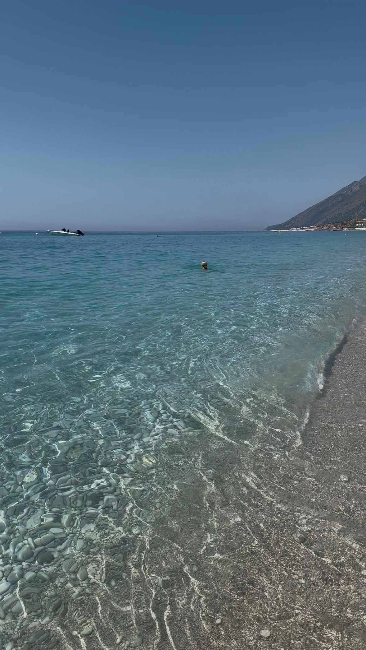 A clear beach with calm water, a distant boat, and a mountain in the background.