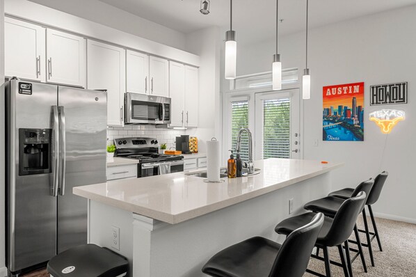 Une cuisine moderne avec des meubles blancs, des appareils en acier inoxydable, un îlot central doté de tabourets et des œuvres d’art décoratives sur les murs.