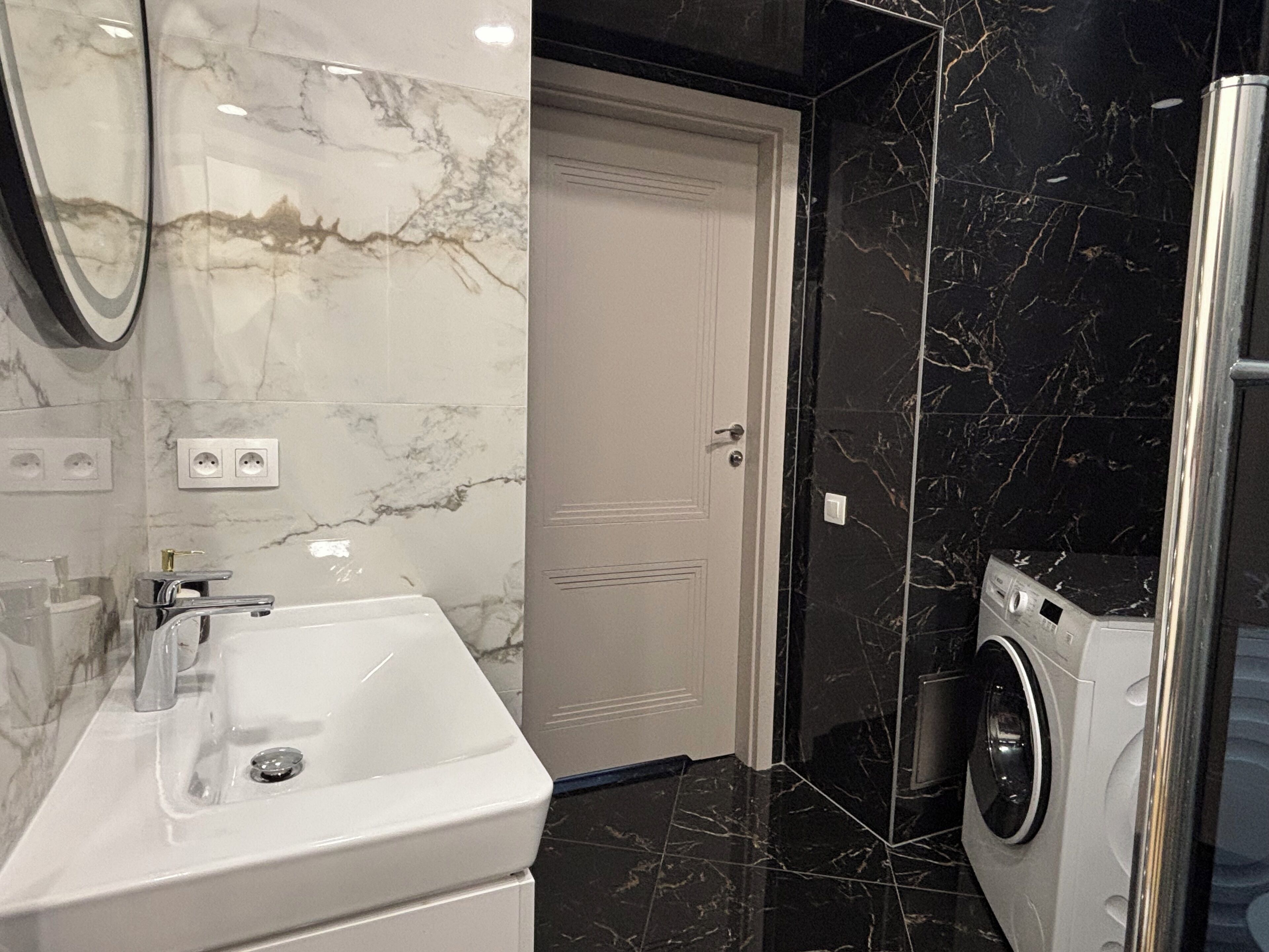 Ein modernes Badezimmer mit einem weißen Waschbecken, einem Spiegel, einer Tür und einer Waschmaschine.