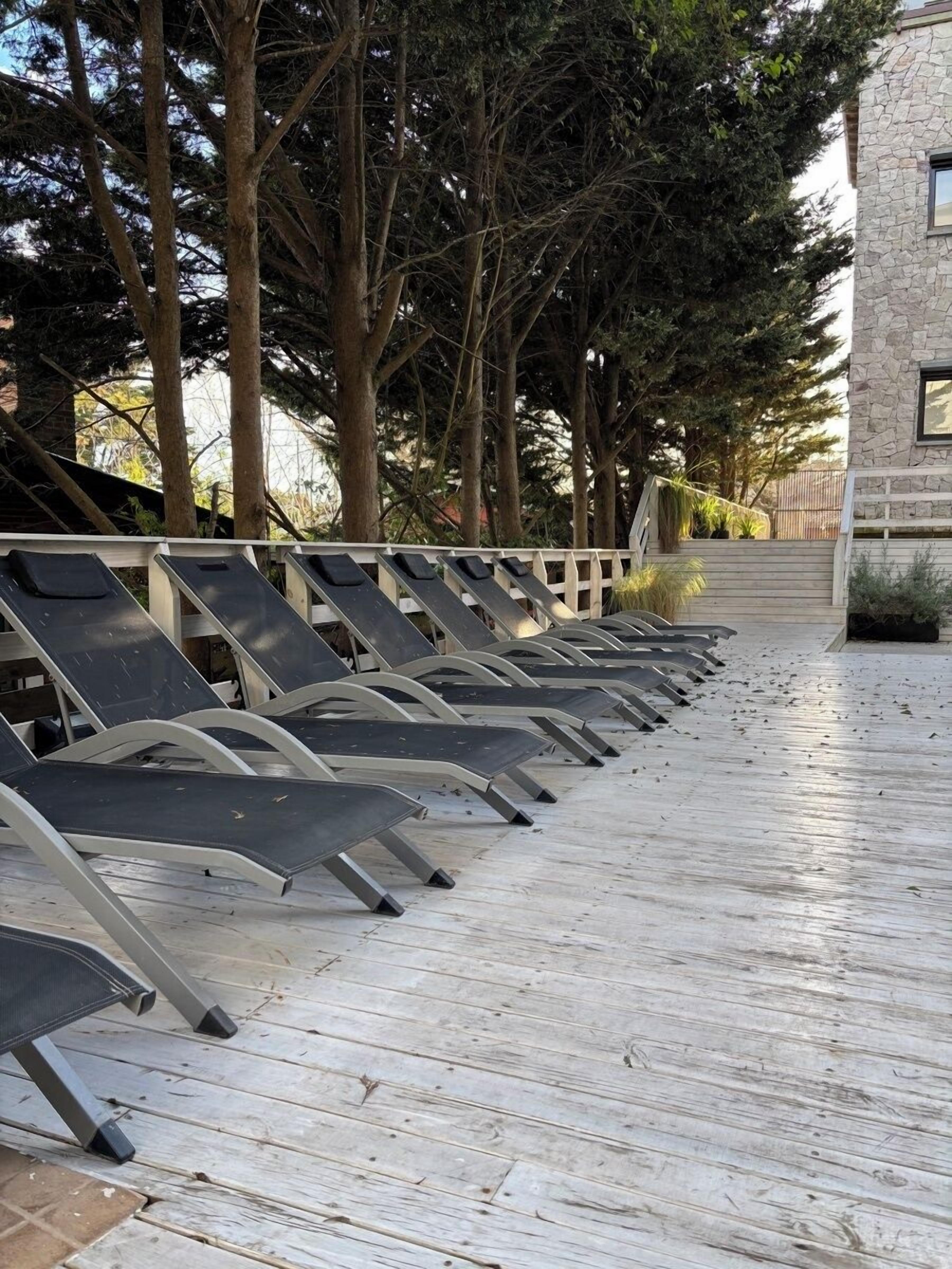 Une rangée de chaises longues disposées sur une terrasse en bois, entourée d’arbres et d’un bâtiment en pierre.
