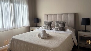 Een net opgemaakt bed met een witte dekbedovertrek, twee kussens met een geometrisch patroon, en een opgerolde handdoek op het bed. Aan beide zijden van het bed staan nachtkastjes met lampen.