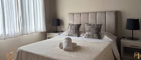 A neatly made bed with a white duvet, two pillows with a geometric pattern, and a rolled towel on the bed. There are two bedside tables with lamps, one on each side of the bed.