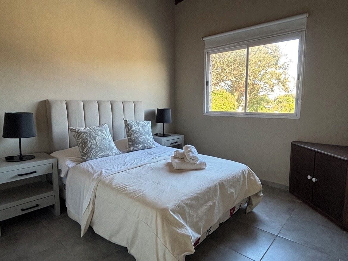A bedroom with a large bed, two bedside tables, a nightstand, a window with a view of trees, and a dark cabinet.