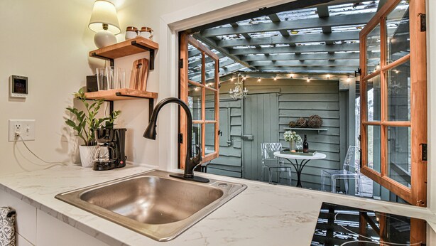 Una cocina con un lavamanos de acero inoxidable, estantes de madera, una cafetera y vistas a un comedor al aire libre a través de amplias ventanas.