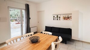 Een eethoek met een houten tafel en witte stoelen, een zwarte bank en een plantenrek.