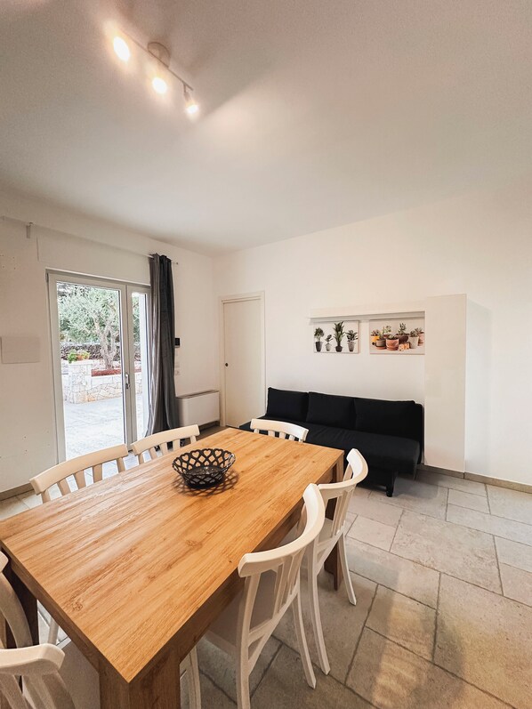 A dining area with a wooden table and white chairs, a black sofa, and a potted plant shelf. - Da Mèmela Trulli e Lamie in the beautiful countryside in Puglia suitable for families (Puglia)