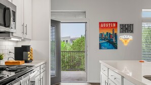 A modern kitchen with white cabinets, a stove, and a coffee maker. There is a balcony with a view of greenery and a cityscape. - Modern 2-bedroom close to Airport & Downtown - Pool, Gym & Balcony (Austin)