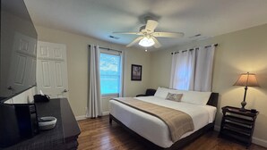 A bedroom with a bed, a ceiling fan, a bedside table with a lamp, a window with curtains, and a door.