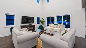 Un salón amplio con televisor de pantalla plana, asientos cómodos, chimenea y ventanas grandes con vistas al exterior.