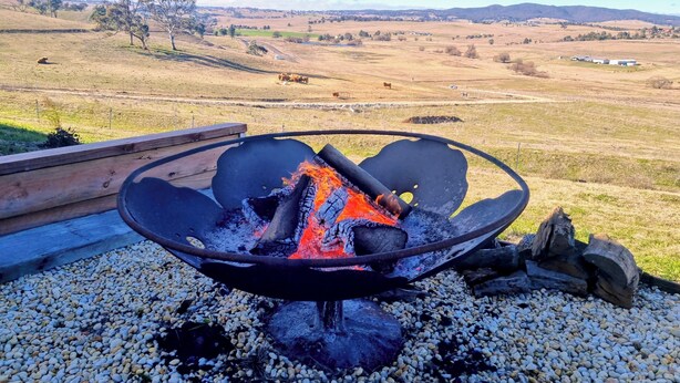 Una fogata con leña, rodeada de guijarros, ubicada en un espacio al aire libre con vistas a un paisaje rural.