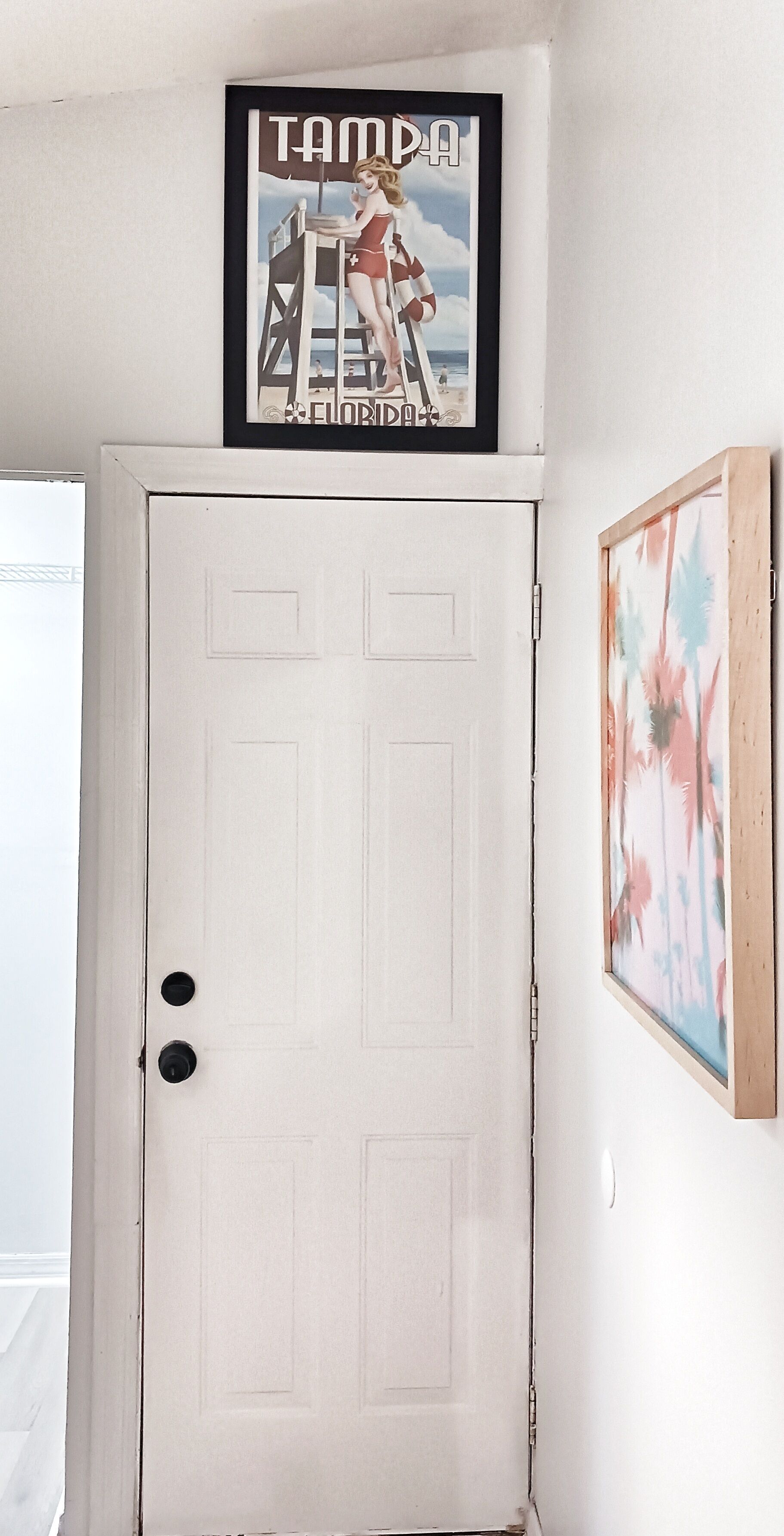 A white door with two round doorknobs, a framed picture of a woman on a ladder above it, and a floral painting to the right.