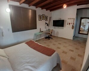 A bedroom with a bed, a desk, a chair, a TV, and a window with blinds.