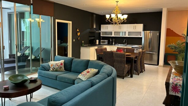 A modern living room with a blue sofa, a dining area with wicker chairs, a kitchen with white cabinets, and a large window. - Delightful 2-bedroom condo with AC, 1/2 Block to 5th Ave and close to the beach! (Playa Del Carmen)