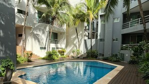 A swimming pool surrounded by a wooden deck, with palm trees and a modern building in the background.