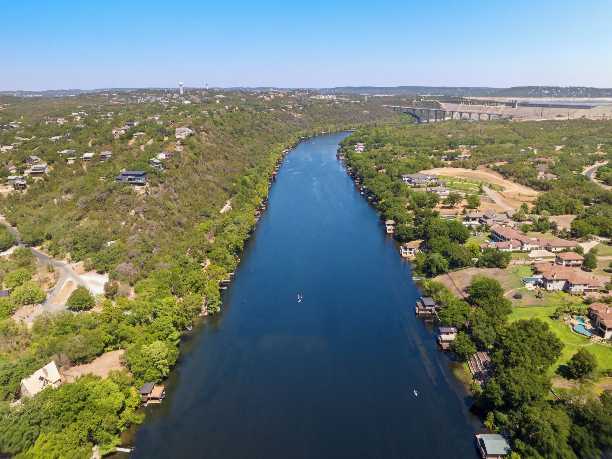 Vista aérea de un río con casas a lo largo de sus orillas, un puente a lo lejos y un cielo despejado.