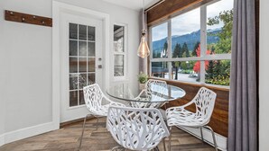 A dining area with a glass table and white chairs, a wooden beam ceiling, and a view of mountains through large windows.