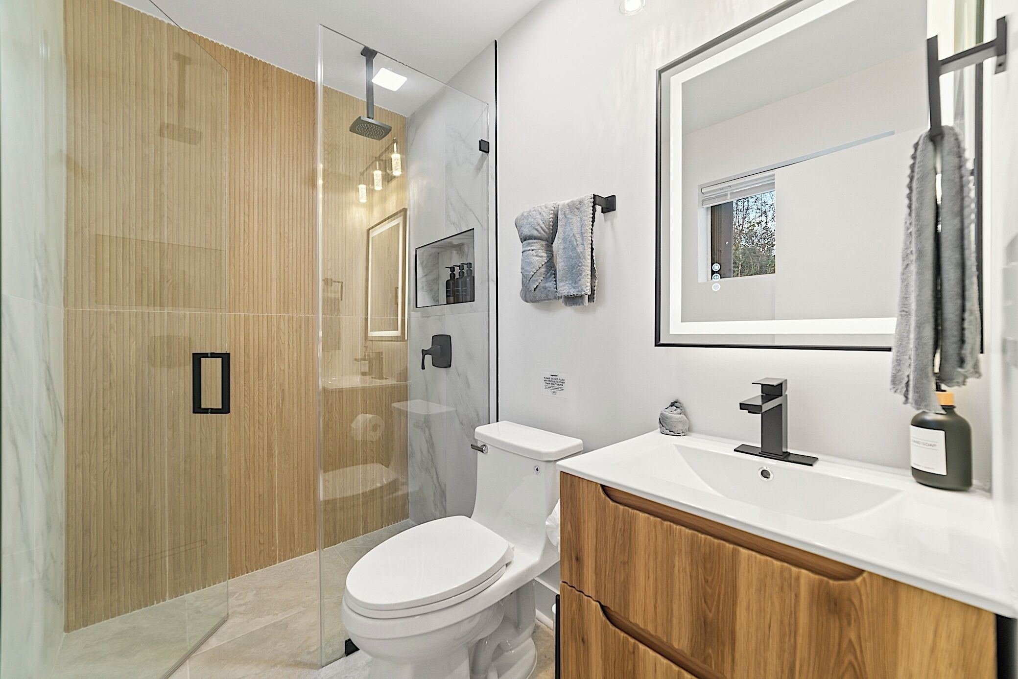 Et moderne badeværelse med bruseniche i glas, et hvidt toilet, et træbord og et stort spejl.