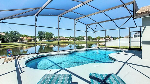 A covered swimming pool with two lounge chairs, a clear view of a lake, and a fenced area. - Smiling Disney Oasis heated pool home in Kissimmee with Game room. (Kissimmee)