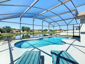 A covered swimming pool with two lounge chairs, a clear view of a lake, and a fenced area. - Smiling Disney Oasis heated pool home in Kissimmee with Game room. (Kissimmee)