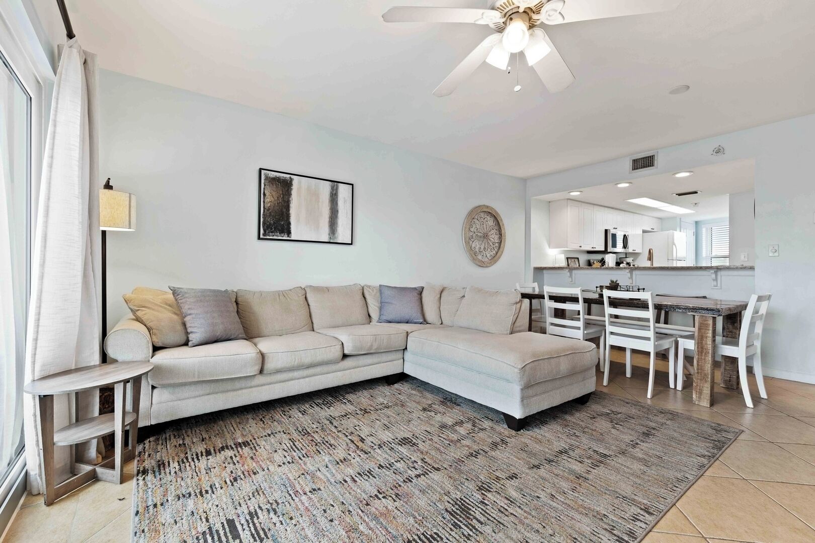 A modern living room with a sectional sofa, a dining table with chairs, a ceiling fan, and a kitchen area in the background.
