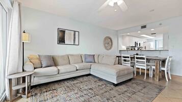 A modern living room with a sectional sofa, a dining table with chairs, a ceiling fan, and a kitchen area in the background.