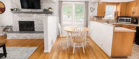 A modern kitchen with a white brick fireplace, a dining area with a wooden table and white chairs, and wooden cabinets.