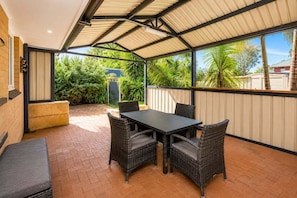Uma área de varanda coberta com mesa, cadeiras e um banco, além de vista para o verde.