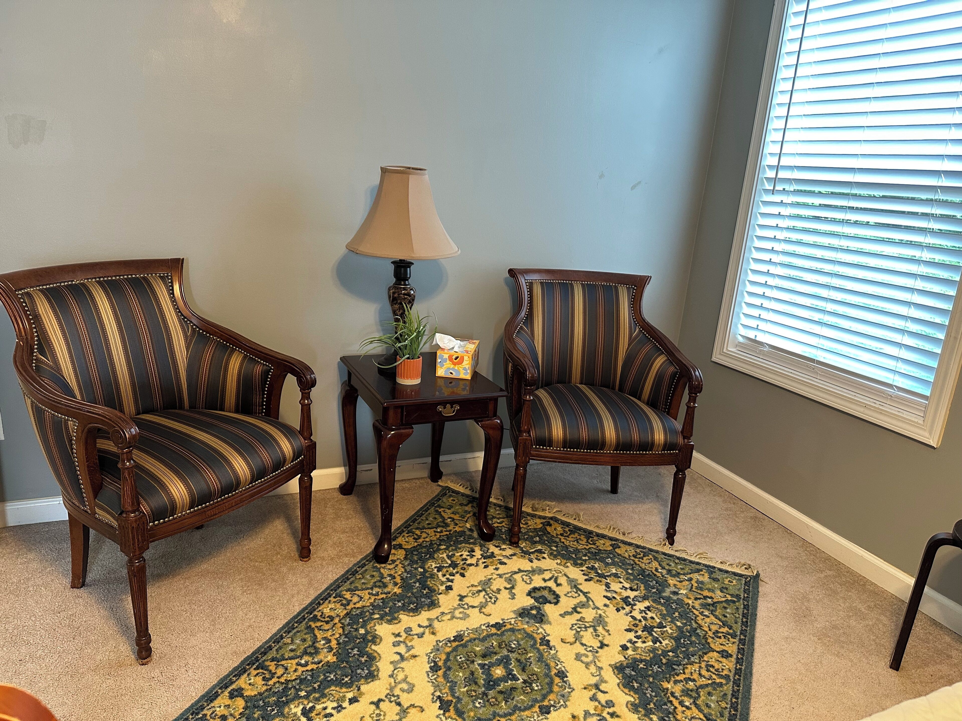 Une pièce avec deux fauteuils à rayures, une petite table avec une lampe, un tapis décoratif et une fenêtre avec des stores.