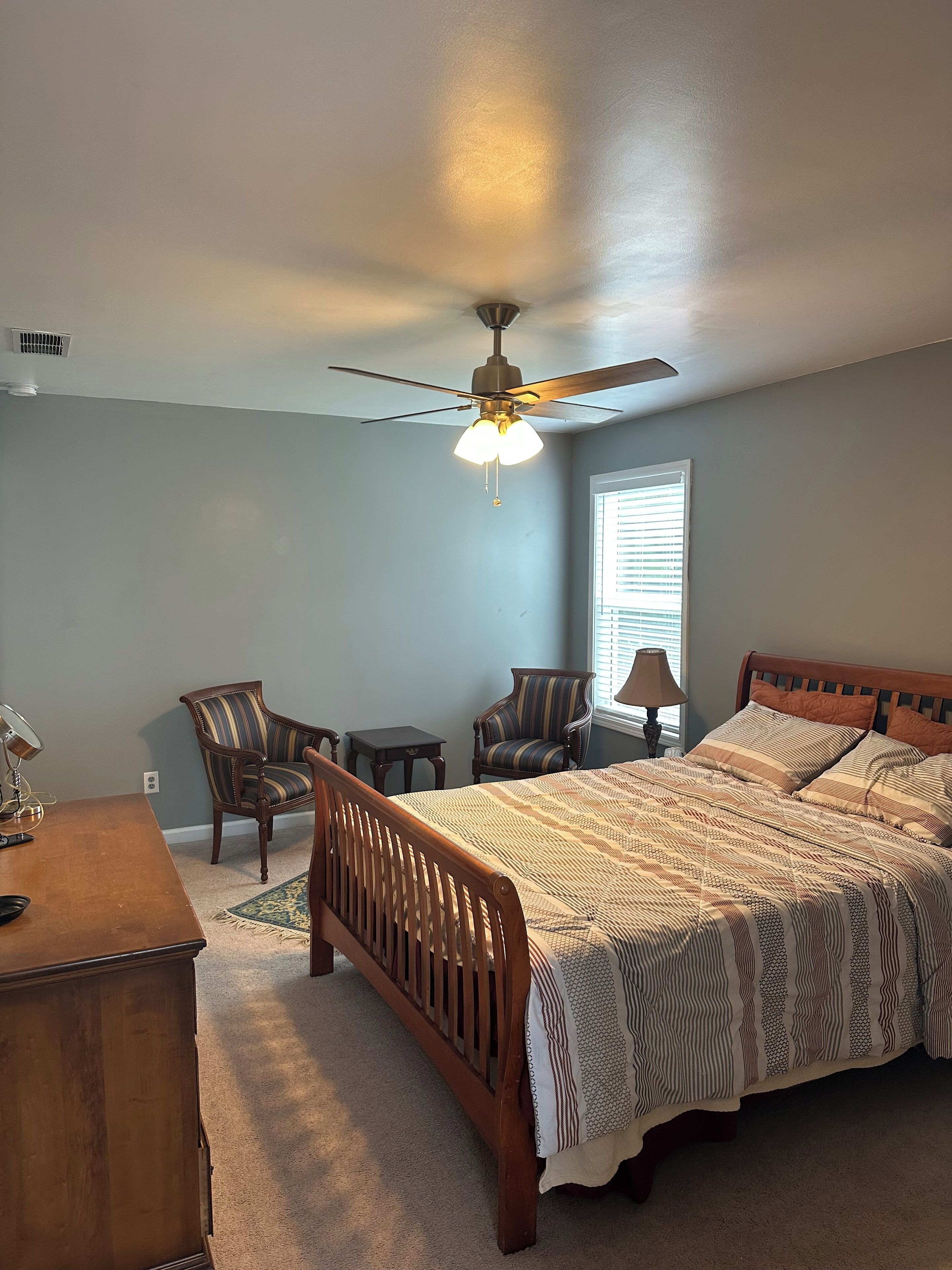A bedroom with a bed, a wooden headboard, a nightstand, a lamp, a chair, a desk, and a ceiling fan.