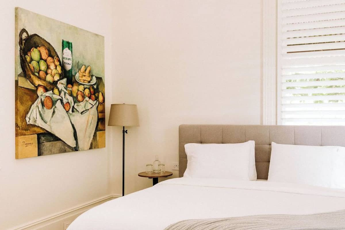 A bedroom with a large bed, a nightstand, a lamp, a painting, and a window with blinds.