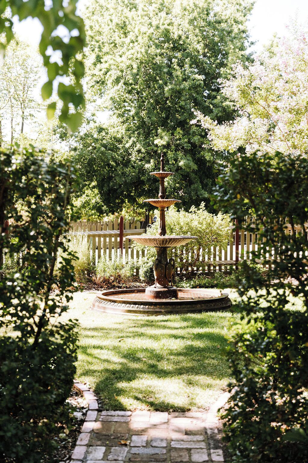Un jardín con una fuente central, rodeado por un sendero de ladrillos y abundante vegetación.